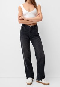 Jeans negros de cintura alta con corte recto, que cuentan con bolsillos frontales y cierre de botón, combinados con una blusa blanca ajustada y zapatillas claras.