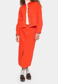 Osoba v jasně oranžové bundě a odpovídající dlouhé sukni s předním rozparkem, bílé roláku, šedých ponožkách a hnědých mokasínech.