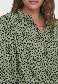 Blusa verde oliva con stampa animale nera, caratterizzata da un scollo a V e un tessuto morbido. Inclusi drappeggi sottili per un tocco di texture e dettaglio.