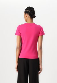 Chemise ajustée à manches courtes en tissu rose vif avec un col rond. Texture lisse, sans motifs ni accents visibles. Portée avec un pantalon noir.