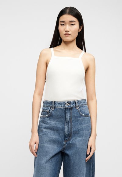 Γυναίκα με μακριά μαύρα μαλλιά που φοράει λευκό τοπ αμάνικο με τετράγωνη λαιμόκοψη και φαρδιά ψηλόμεσα μπλε τζιν, στέκεται μπροστά σε απλό φόντο.