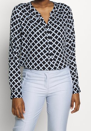 Vrouw draagt een zwart blouse met lange mouwen met blauw-wit ruitpatroon en lichtblauwe aansluitende broek, staande tegen een effen achtergrond.
