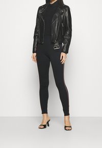 Zwarte leren motorjack met zilveren accenten, gedragen over een strak zwart topje en hooggegarande zwarte leggings met rode zijstrepen en zwarte hakken.