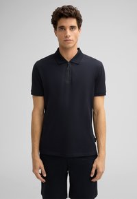 Czarny polo z krótkim rękawem, zapinany na zamek, o gładkiej teksturze. Noszony z ciemnymi szortami, na neutralnym tle.