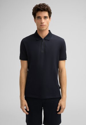 Czarny polo z krótkim rękawem, zapinany na zamek, o gładkiej teksturze. Noszony z ciemnymi szortami, na neutralnym tle.