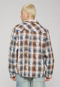 Camicia a maniche lunghe a quadri in tonalità di marrone, blu navy e bianco, caratterizzata da un motivo decorativo e polsini con bottoni. Vestibilità comoda e collo a punta.