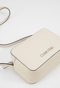 Słomkowa skórzana torebka w kształcie prostokąta, z logo ze srebrnym wykończeniem i kontrastującymi ciemnymi stębnowaniami. Zawiera pasek na ramię.