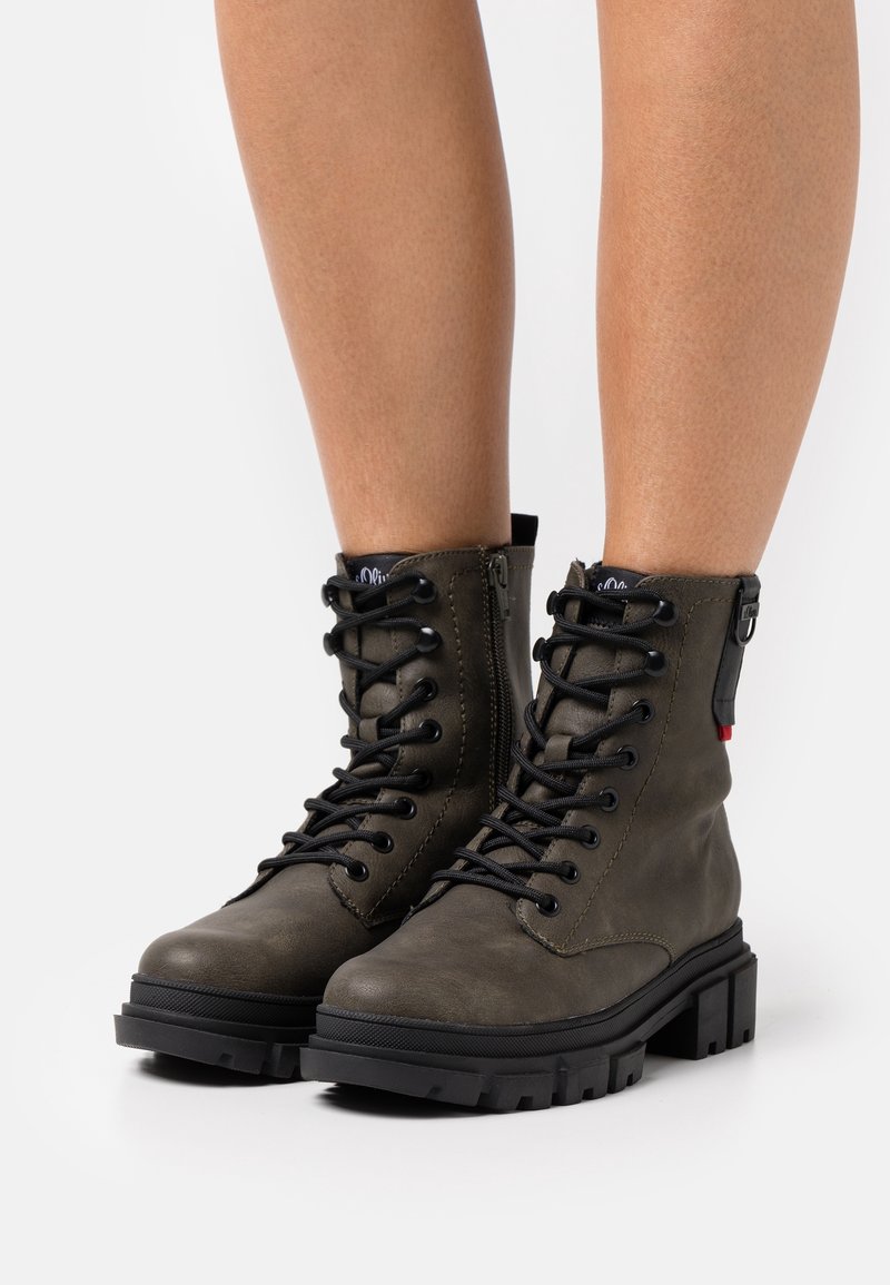 Une paire de bottes de combat vert olive à lacets avec des semelles noires épaisses, portées sur des jambes, sur fond blanc.