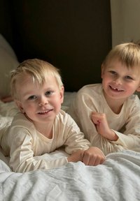 Kaksi lasta vaaleanvaaleassa, kuviollisessa, pitkähihaisessa pyjamassa makaavat valkoisella sängyn päällisellä hymyillen kameraan.