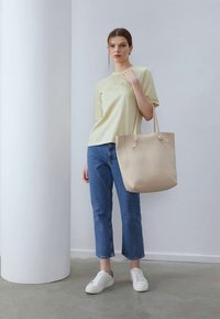 Sac bej cu mânere scurte, textură netedă. Purtați cu un tricou verde deschis, blugi croiți albaștri și adidași albi pe o podea simplă.