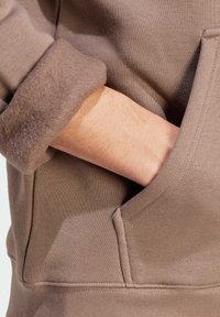 Ruka částečně uvnitř přední kapsy hnědého mikiny s fleecovou podšívkou a vyhrnutým rukávem.