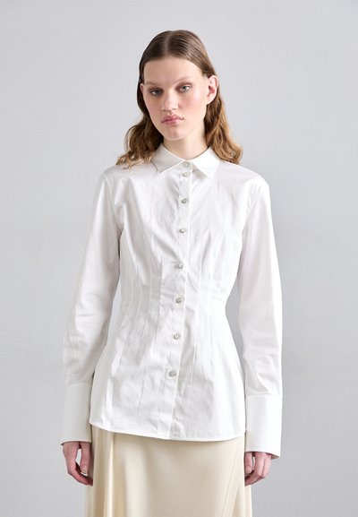 Chemise blanche ajustée avec un col pointu, des manches longues et une fermeture à boutons. Tissu en coton lisse avec des coutures verticales pour la silhouette.