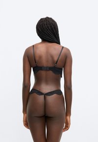 Σετ εσώρουχα από μαύρη δαντέλα, που �περιλαμβάνει σουτιέν με λεπτές τιράντες και κούμπωμα με αγκράφα, καθώς και τόνγκ με κυματιστά άκρα και ελάχιστη κάλυψη.