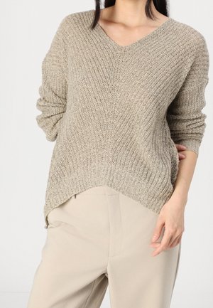 Femme portant un pull en maille texturé beige à manches longues et un pantalon beige clair taille haute, debout devant un fond blanc.