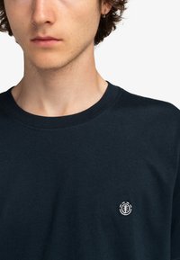 Námořnická modrá bavlněná tričko s kulatým výstřihem, zdobené malým bílým logem na levé straně hrudi. Měkká textura, klasický střih.