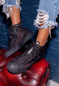 Svarta hög-top sneakers med mesh ovandel, gummisula och platta snören. Bärs med slitna uppvikta blå jeans på en röd läderyta.