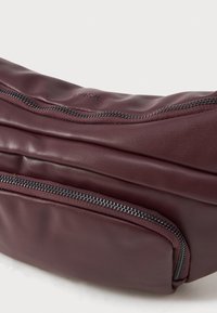 Sac en cuir bordeaux avec une texture lisse ; équipé d'une fermeture éclair supérieure et d'une poche avant avec une fermeture éclair métallique contrastante. Design minimaliste.
