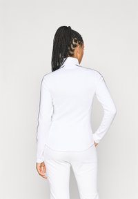 Polo shirt bianco a maniche lunghe con colletto alla coreana, caratterizzato da strisce laterali nere e un design aderente. Tessuto liscio e materiale leggero.