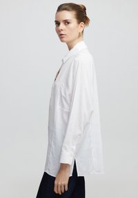 Camicia bianca con bottoni, realizzata in tessuto leggero, caratterizzata da un colletto classico, maniche lunghe, spacchi laterali e dettagli testurizzati discreti.