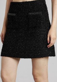 Minifalda negra texturizada con dos bolsillos frontales adornados con ribete negro. El material tiene un acabado difuso y brillante, lo que añade dimensión al diseño.