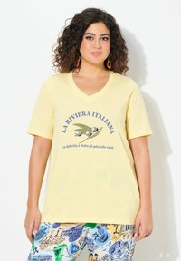 Žluté tričko s výstřihem do V a krátkými rukávy, na kterém je grafický potisk oliv a text, spárované s kalhotami s barevným vzorem.