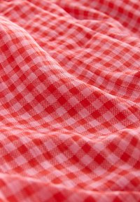 Červeno-růžová gingham textilie s kostkovaným vzorem, s měkkou texturou a lehkým provedením, zavěšená a mírně pomačkaná.