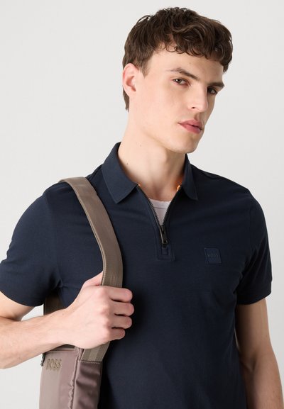 Polo bleu marine avec un col zippé et un petit logo. Le modèle tient un sac beige clair avec des anses en tissu, montrant un matériau lisse et des lignes épurées.
