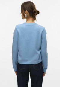 Vrouw met bruin haar in een knot, die zilveren oorringen draagt, een lichtblauwe trui en donkerblauwe jeans, van achteren afgebeeld op een witte achtergrond.