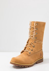 Botte d'hiver marron clair à lacets avec doublure en fourrure, œillets métalliques, semelle en caoutchouc et logo en relief sur le talon, présentée sur un fond blanc uni.
