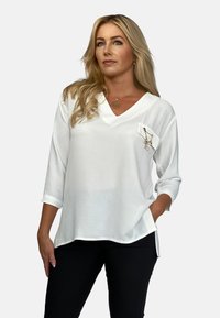 Biała bluzka z dekoltem w serek i rękawami o długości 3/4, z kieszonką i dekoracyjnymi elementami. Teksturowana tkanina z bocznymi rozcięciami dla wygody.