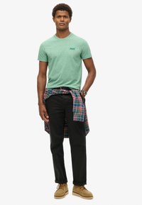 Green heathered t-shirt with a small logo, black pants, and a plaid shirt tied at the waist. Tan boots complete the outfit.