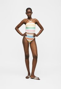 Γυναίκα που στέκεται με τα χέρια στη μέση, φορώντας πολύχρωμο ριγέ μαγιό με λαιμόκοψη halter και αντίστοιχα σανδάλια σε μονόχρωμο φόντο.