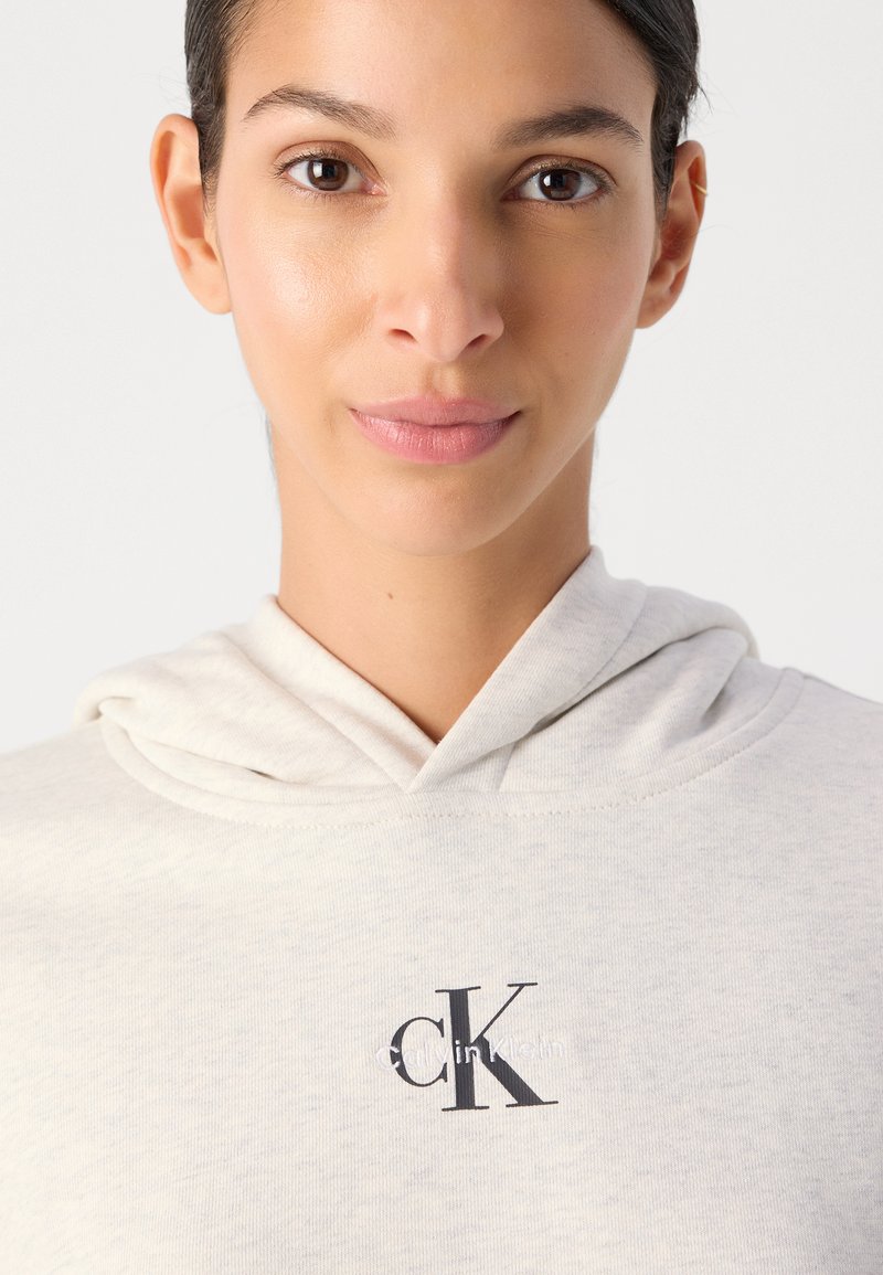 Lichtgrijze hoodie met een zachte textuur en een trekkoord, voorzien van een opvallend zwart "CK"-logo op de borst. Eenvoudig en casual ontwerp.