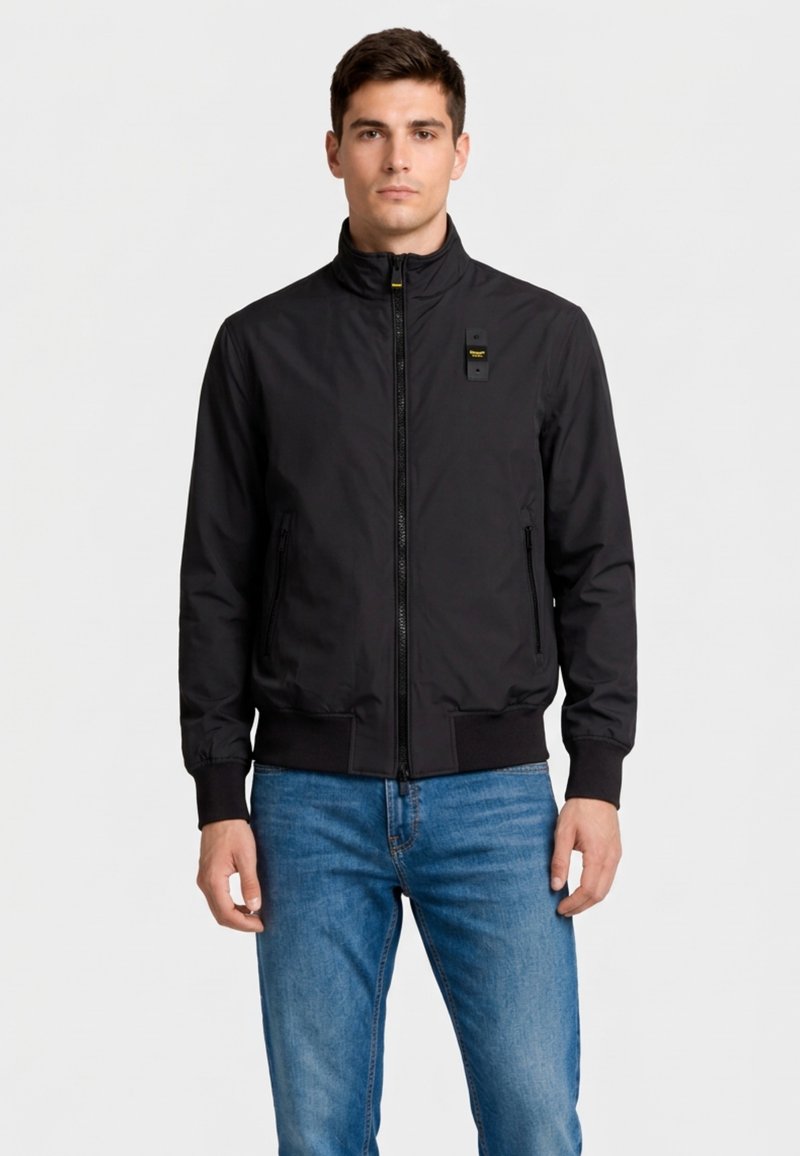 Uomo che indossa una giacca nera con zip, polsini e orlo a coste, in piedi davanti a uno sfondo bianco uniforme, vestito con jeans blu.