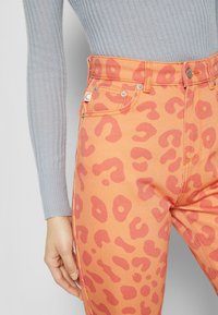 Jean en denim orange avec imprimé léopard rose. Comprend cinq poches, des accessoires en argent et une passants de ceinture à la taille haute. Tissu texturé.