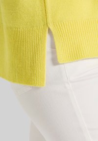 Gelber gestrickter Pullover mit geripptem Saum und seitlichen Schlitzen, kombiniert mit weißen Hosen, die eine strukturierte Stoffqualität und einen lebendigen Farbkontrast zeigen.