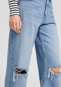 Ingrandimento di una persona che indossa jeans strappati blu chiaro e una maglietta a maniche lunghe bianca e nera a righe, con una mano rilassata di fianco.