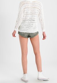 Osoba oblečená v bílém pleteném topu s dlouhým rukávem, zelených kraťasech s ohrnutými nohavicemi a bílých teniskách s vysokým svrškem, stojící zády k bílé pozadí.