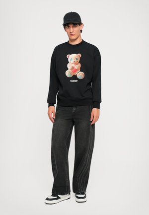 Felpa nera con una grafica colorata di un orsetto, abbinata a jeans neri larghi. Il modello indossa un cappellino da baseball e sneakers.