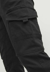 Pantalons cargo noirs avec plusieurs poches, plis cousus et finition texturée. Le tissu semble durable, adapté à un usage pratique.