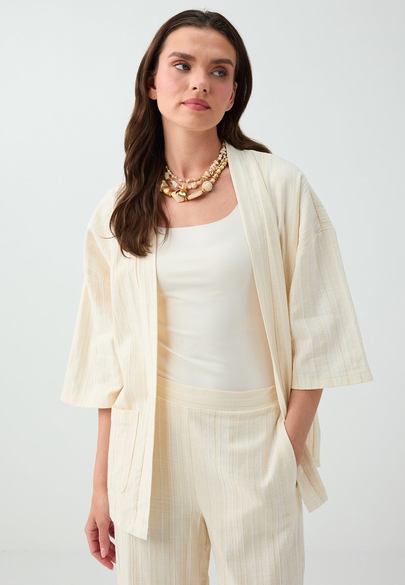 Femme aux longs cheveux foncés portant une veste ouverte crème côtelée, un pantalon assorti, un haut blanc et des colliers superposés en or et perles.
