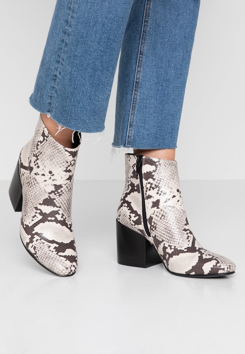 Personne portant des bottines à motif peau de serpent avec talons carrés et fermetures éclair latérales, associées à un jean bleu raccourci.