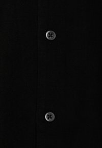 Chemise noire à boutons avec un tissu texturé, dotée de deux boutons noirs et d'un design de patte de boutonnage verticale. Esthétique simple et minimaliste.