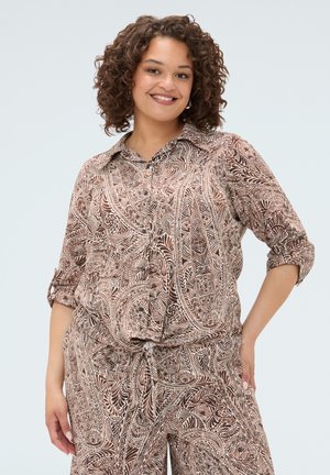 Donna sorridente con capelli ricci che indossa una camicia marrone e beige con motivo paisley abbottonata e pantaloni coordinati su sfondo neutro.