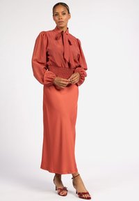 Rustfarbene Bluse mit einer Schleife, strukturiertem Stoff, Puffärmeln, kombiniert mit einem passenden langen Rock. Rote Sandalen mit Riemchen und Absatz.