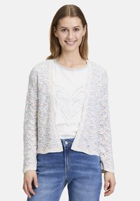 Virkat cardigan med en vågig mönster i pastellfärger, öppen front och långa ärmar. I kombination med en vit skjorta och blå jeans.