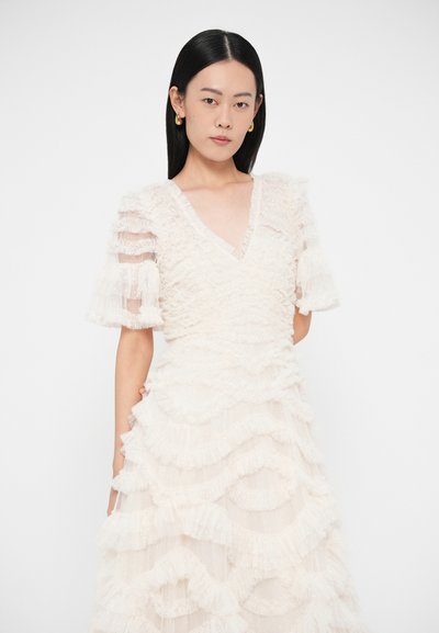 Młoda kobieta z prostymi czarnymi włosami, nosząca złote kolczyki koła oraz białą sukienkę o fakturze, z warstwowymi falbanami i przezroczystymi bufkami na rękawach.