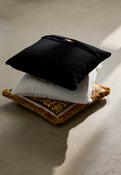 Tre staplade dekorativa kuddar: en svart plyschkudde, en vit kudde med ett utsmyckat mönster, och en fransad guld- och mönstrad kudde.