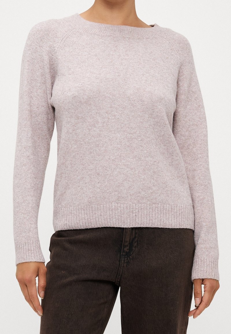 Femme portant un pull en maille gris clair à manches longues associé à un pantalon marron foncé, debout devant un fond uni.