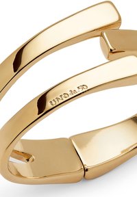 Bracelet jonc en ton or avec une surface lisse et polie. Présente un design moderne avec des formes courbes entrelacées et un détail de logo subtil.